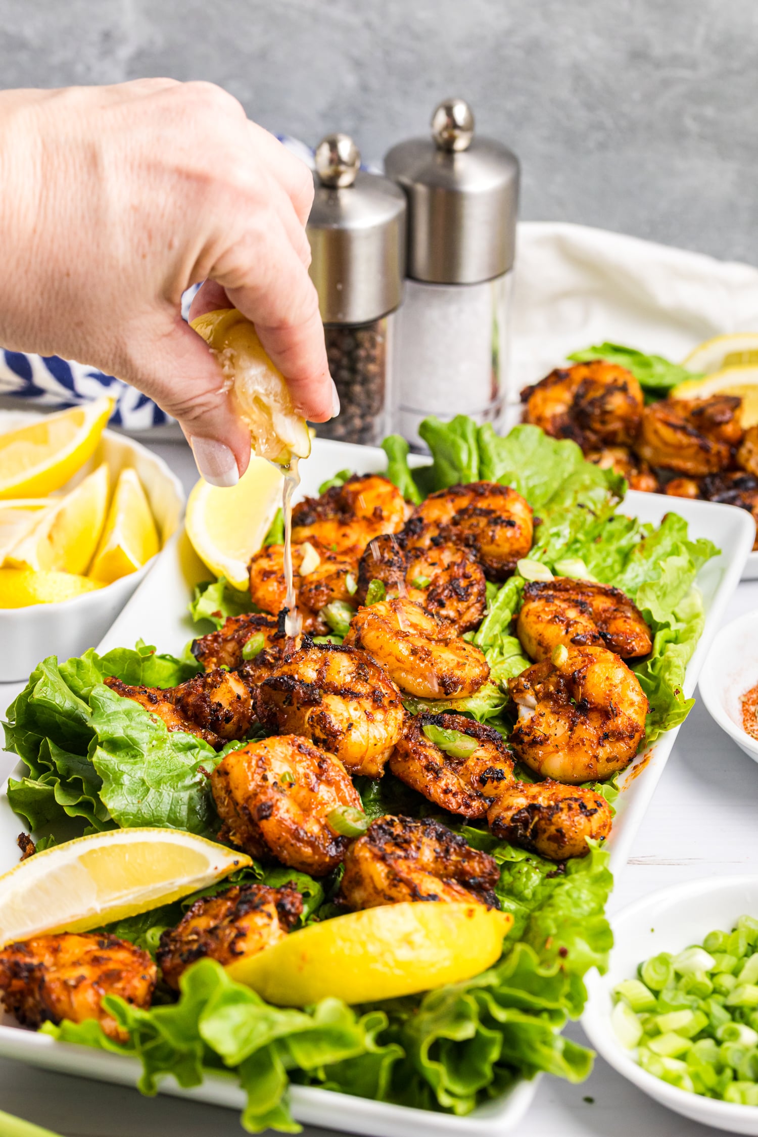 Hand squeezing lemon juice over grilled shrimp served on lettuce with lemon wedges and green onions.