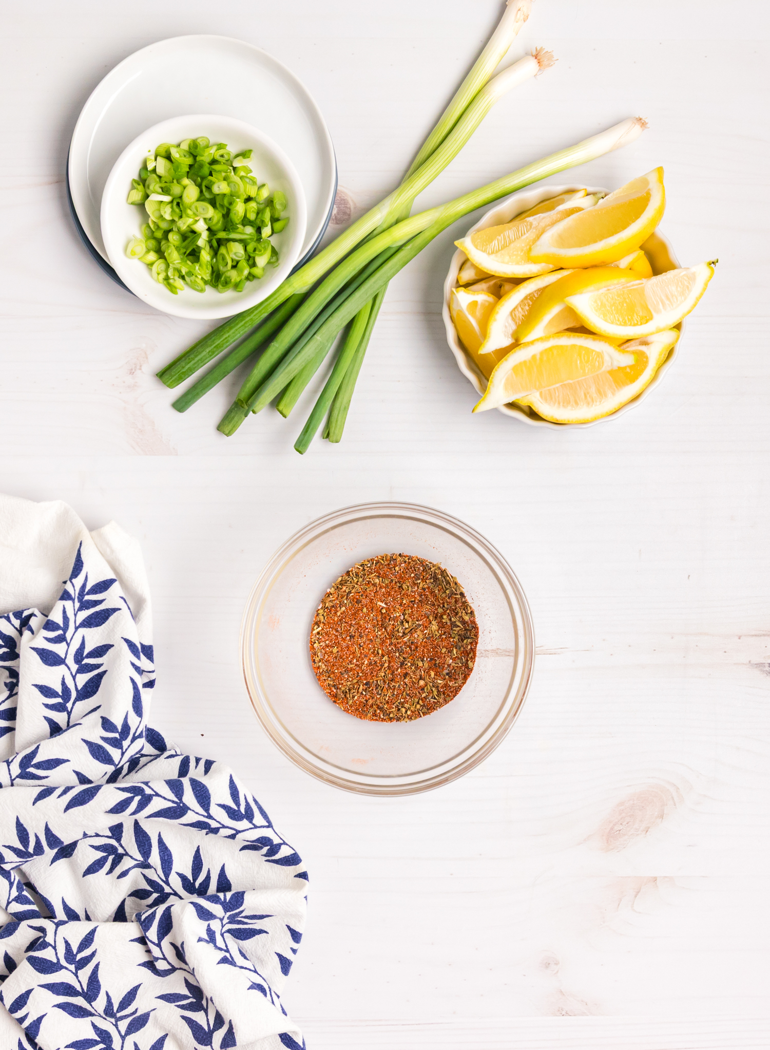 Chopped green onions, whole scallions, lemon wedges, and a bowl of spice blend arranged on a white surface.