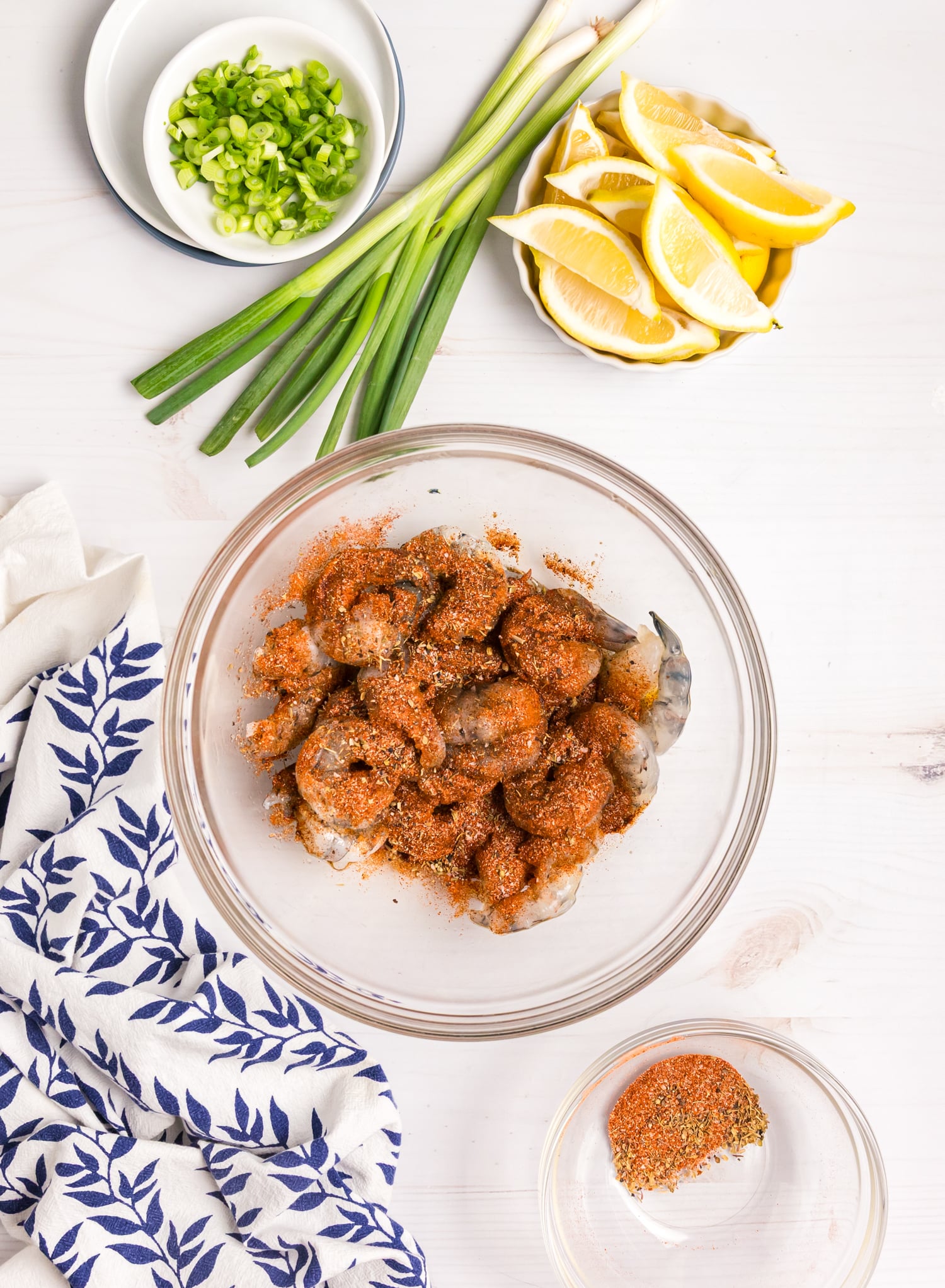 Raw shrimp in a glass bowl coated with a reddish spice mixture, with green onions and lemon wedges nearby.