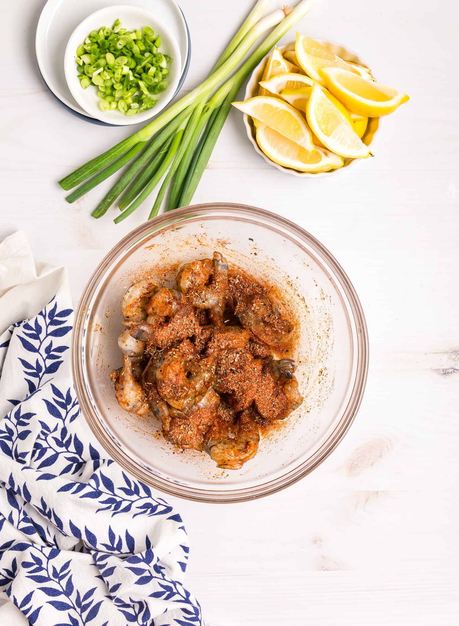 Seasoned raw shrimp in a bowl, coated evenly with spices, surrounded by fresh scallions and lemon slices.