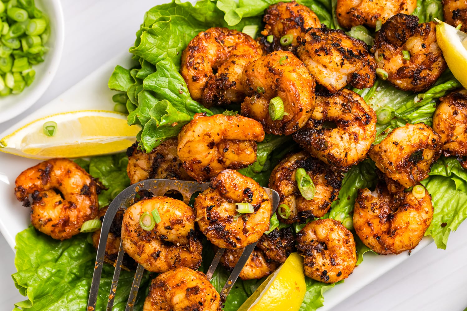 Close-up of grilled blackened shrimp on a bed of lettuce, garnished with sliced green onions and lemon wedges, with a fork lifting shrimp.