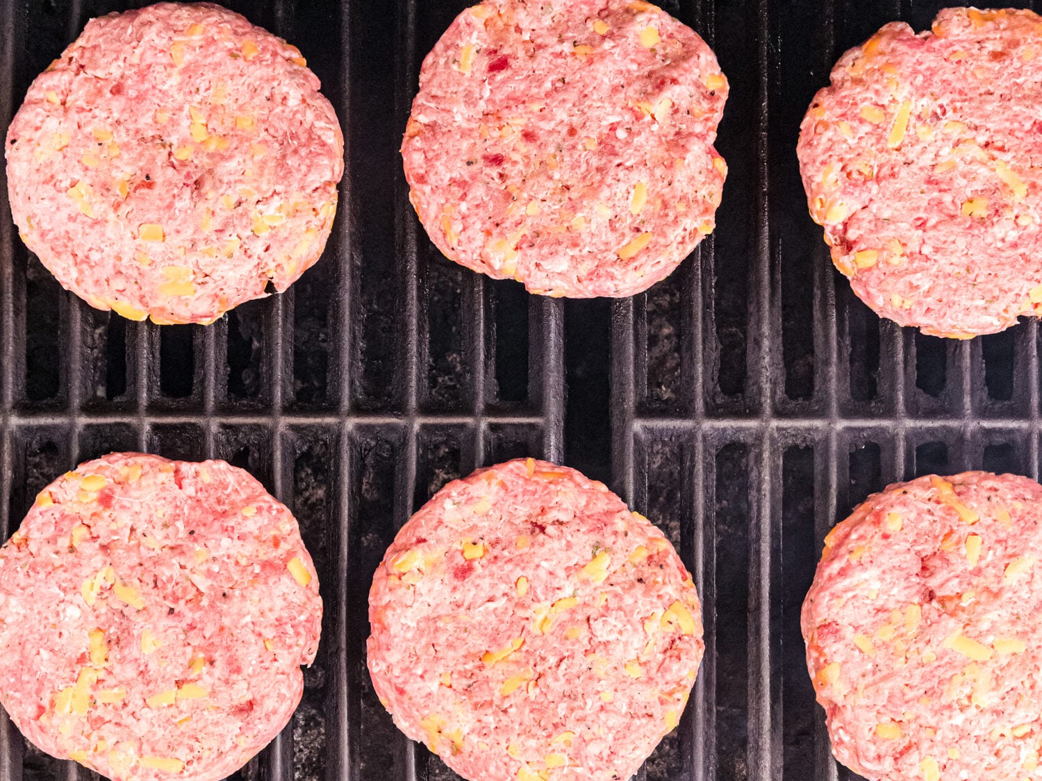 Several raw burger patties cooking on a grill, evenly spaced across the grates.