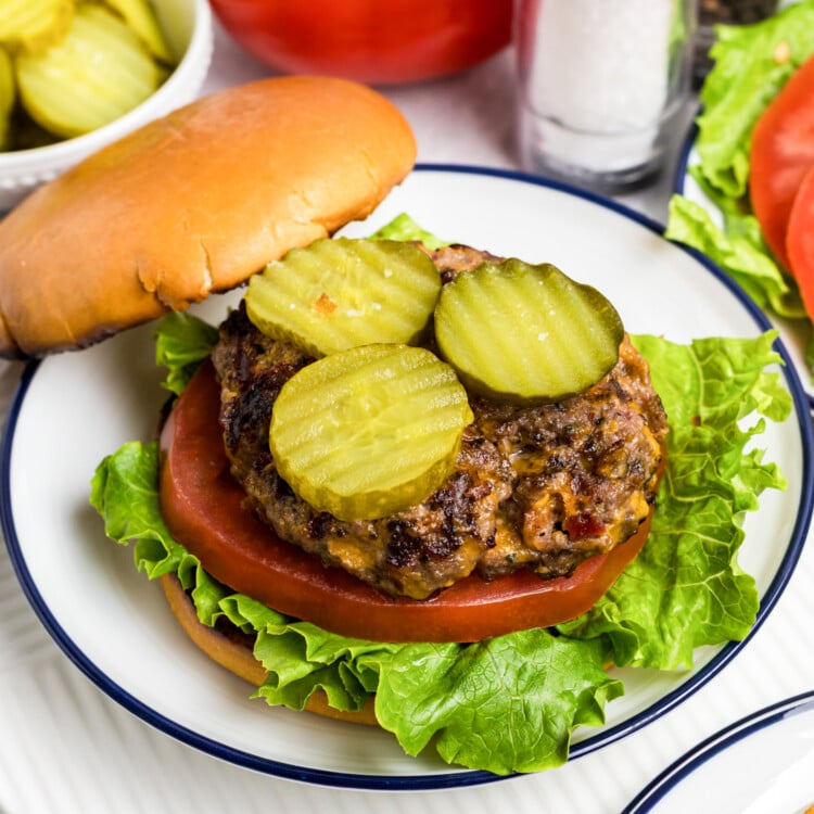 Close-up of a juicy cheeseburger on a bun, topped with sliced pickles, tomato, and crisp lettuce, served on a plate with whole tomatoes, pickles, and salt and pepper shakers in the background.