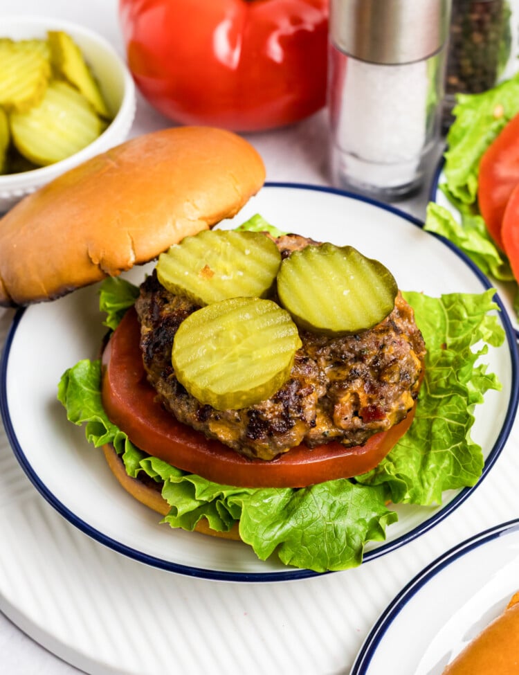 Close-up of a juicy cheeseburger on a bun, topped with sliced pickles, tomato, and crisp lettuce, served on a plate with whole tomatoes, pickles, and salt and pepper shakers in the background.
