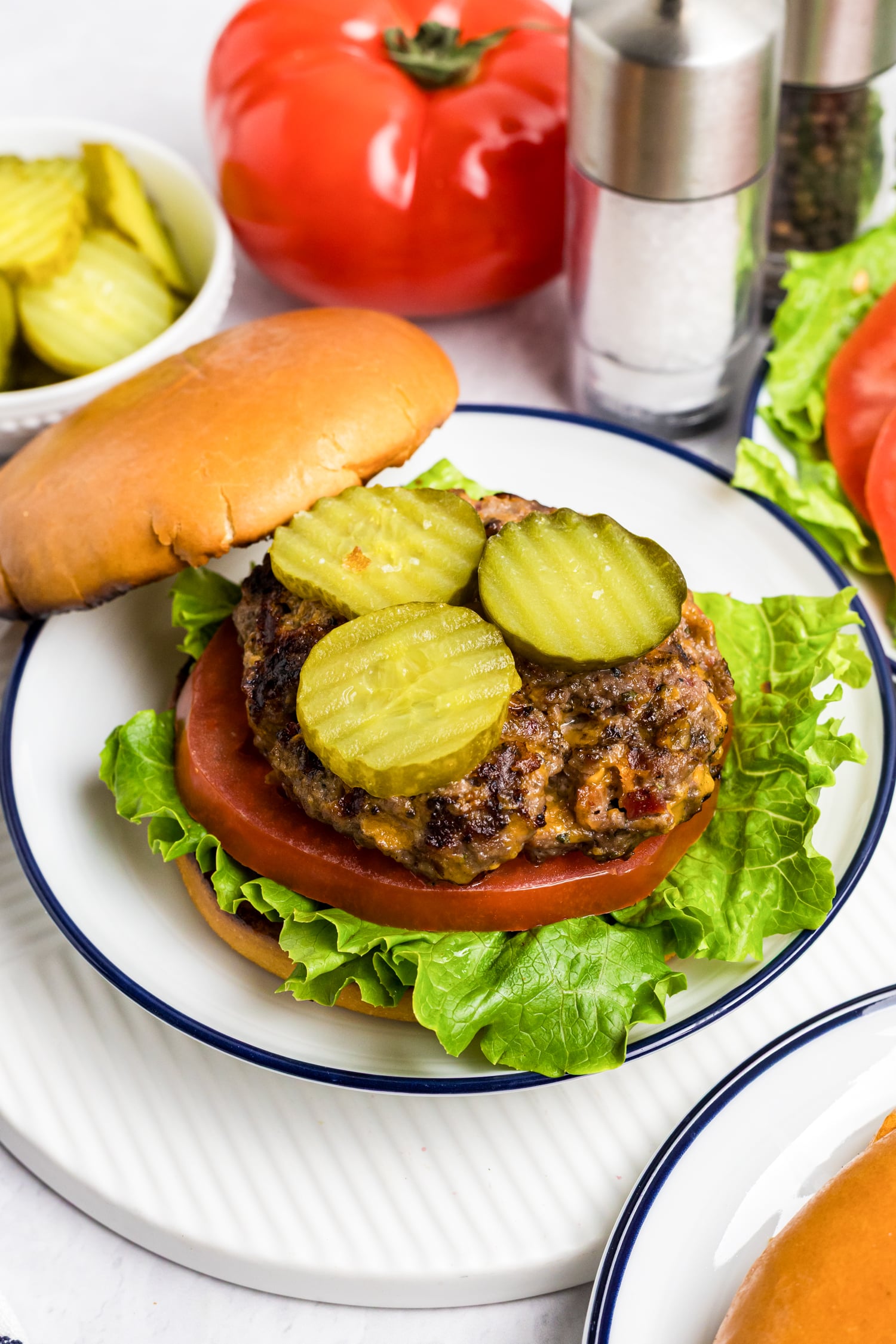 Close-up of a juicy cheeseburger on a bun, topped with sliced pickles, tomato, and crisp lettuce, served on a plate with whole tomatoes, pickles, and salt and pepper shakers in the background.