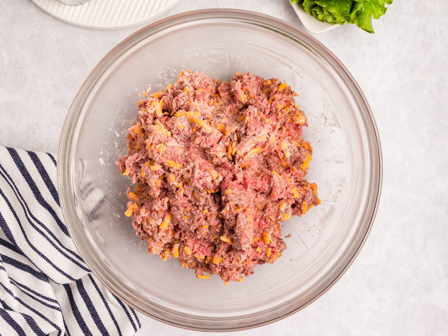 A bowl of raw ground beef mixed with shredded cheese beside sliced tomatoes, lettuce, and pickles on a plate.