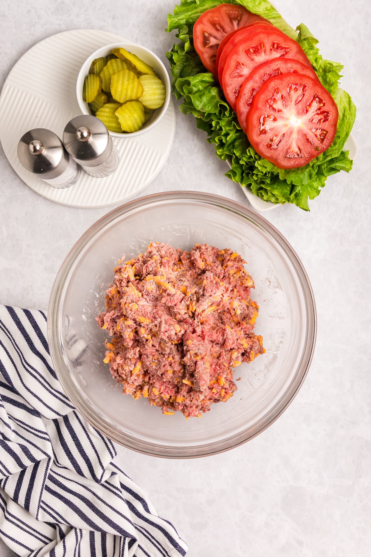 A bowl of raw ground beef mixed with shredded cheese beside sliced tomatoes, lettuce, and pickles on a plate.