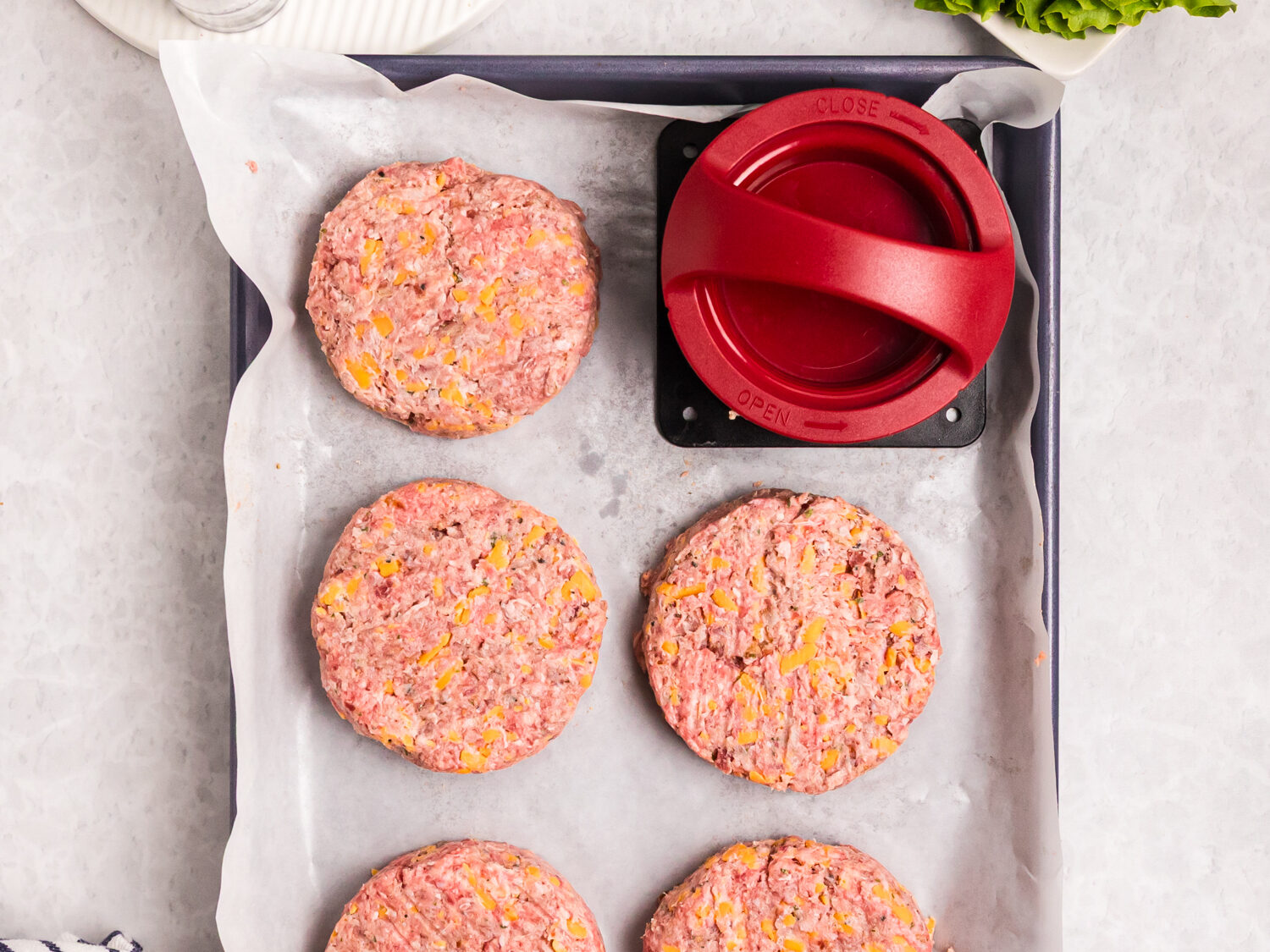 Raw burger patties mixed with cheese arranged on a parchment-lined baking sheet next to a burger press and fresh toppings.