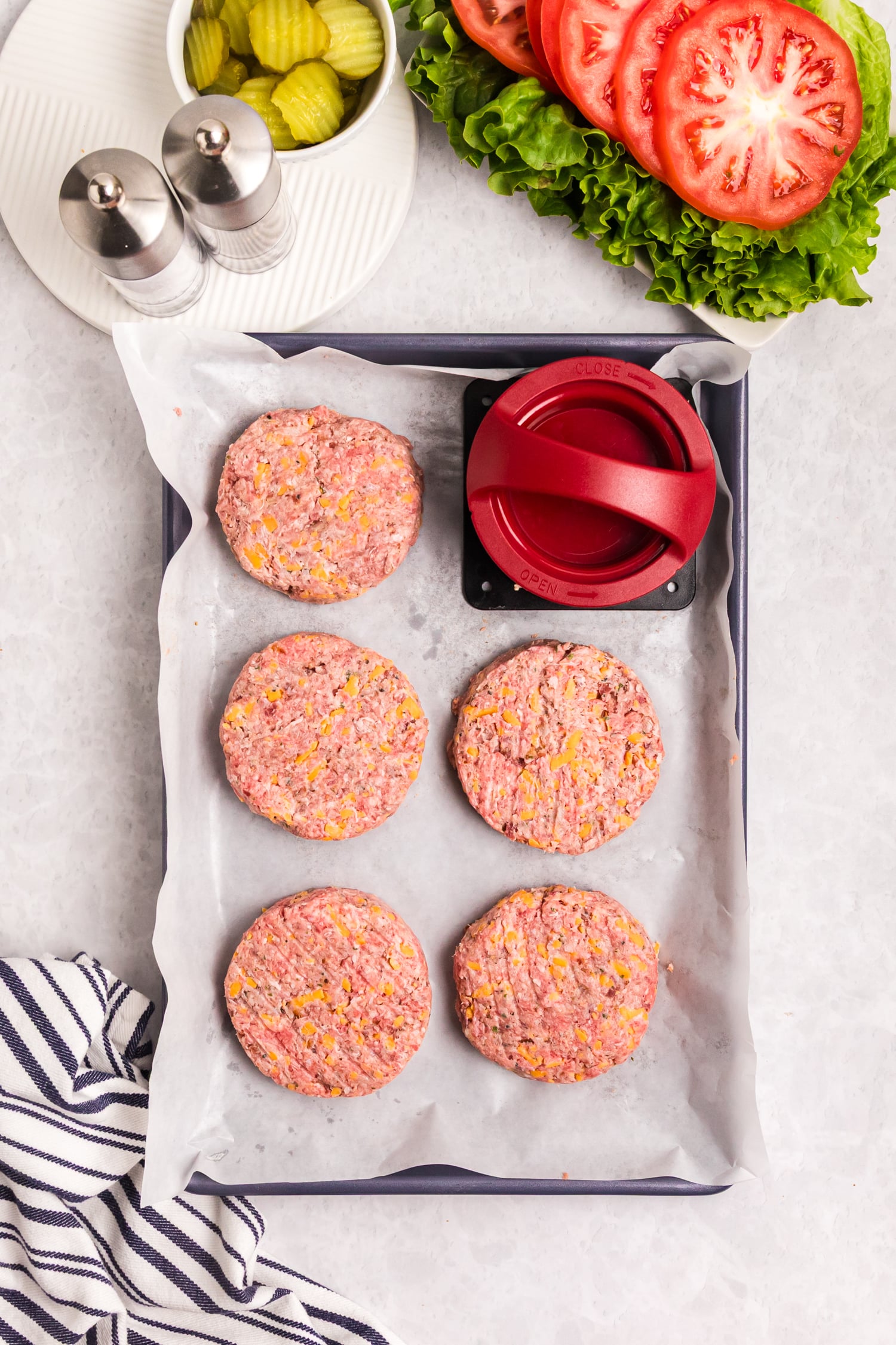 Raw burger patties mixed with cheese arranged on a parchment-lined baking sheet next to a burger press and fresh toppings.