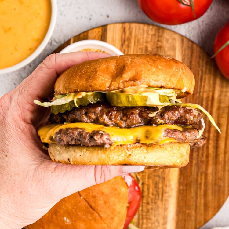 Hand holding a double cheeseburger with pickles, lettuce, and sauce on a toasted bun.