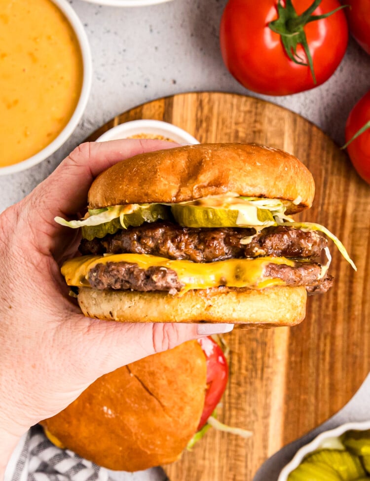 Hand holding a double cheeseburger with pickles, lettuce, and sauce on a toasted bun.