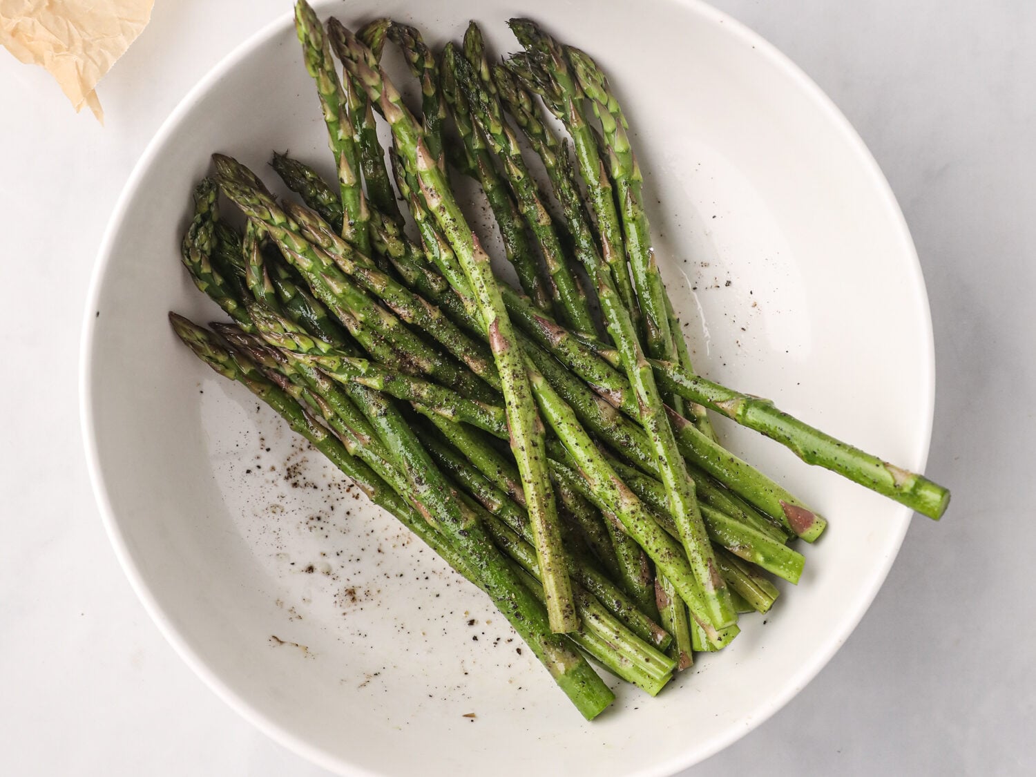 Fresh asparagus spears seasoned with salt and pepper in a white bowl, with slices of raw bacon visible on parchment paper beside it.