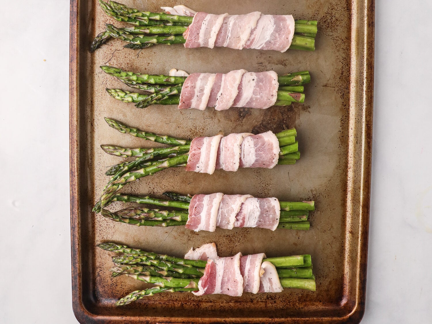 Bundles of asparagus spears wrapped in raw bacon arranged evenly on a baking sheet, ready for grilling.