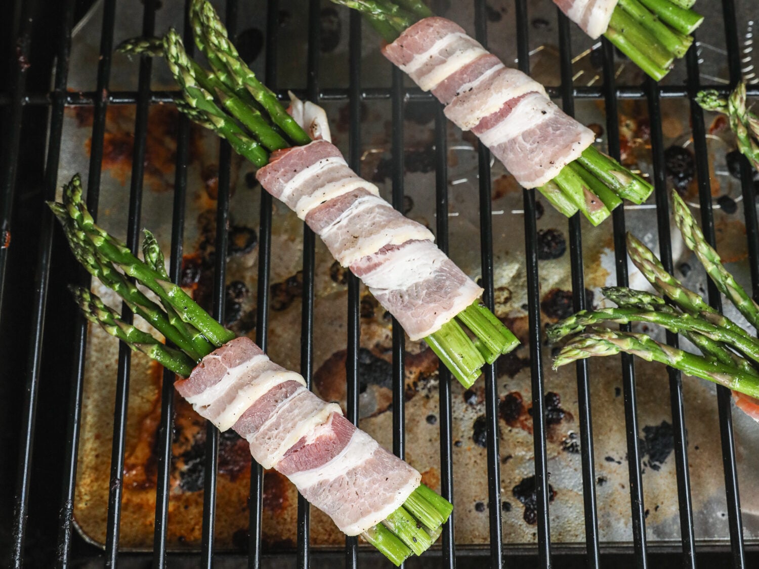 Asparagus bundles wrapped in raw bacon placed on a grill grate above a drip tray, beginning the cooking process.