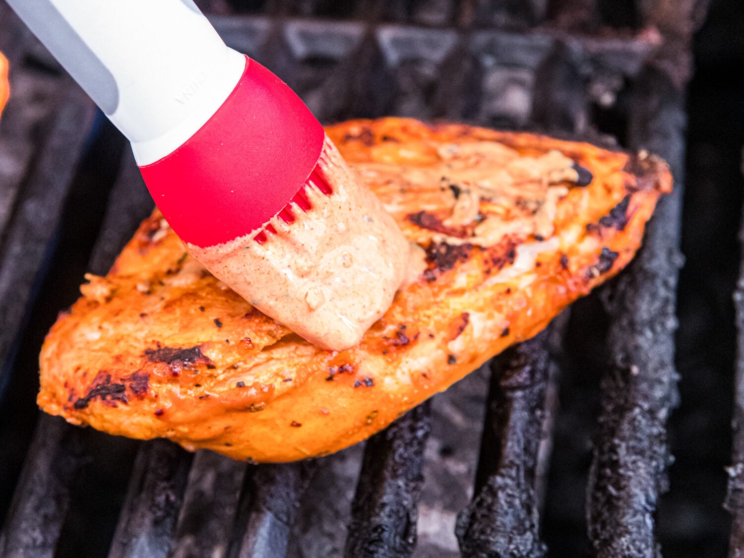 Silicone basting brush spreading marinade over grilled chicken breasts as they cook on an outdoor grill.