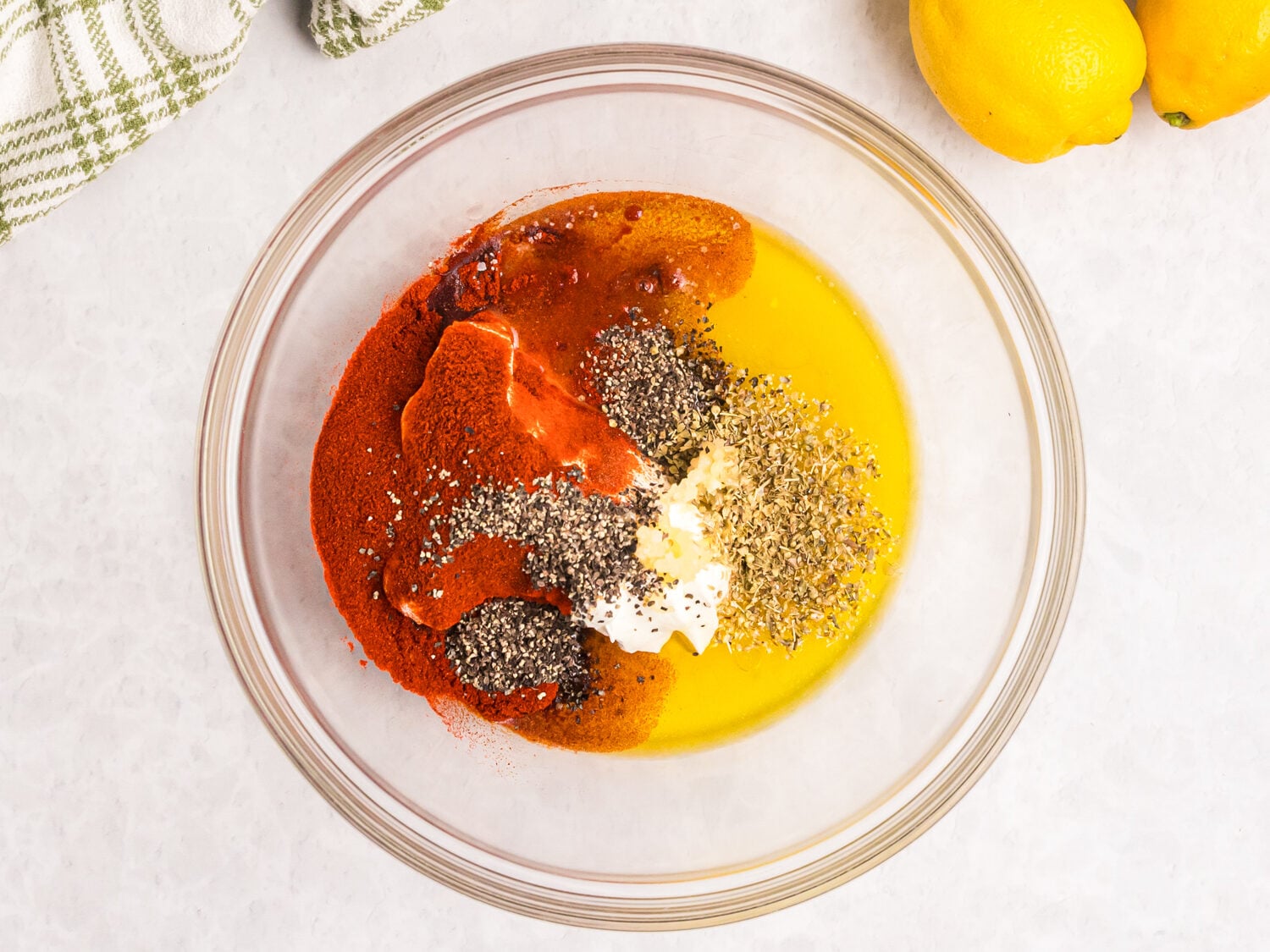 Glass mixing bowl filled with olive oil, yogurt, paprika, herbs, and seasonings for Mediterranean chicken marinade, surrounded by lemons, olive oil, and salt and pepper shakers.