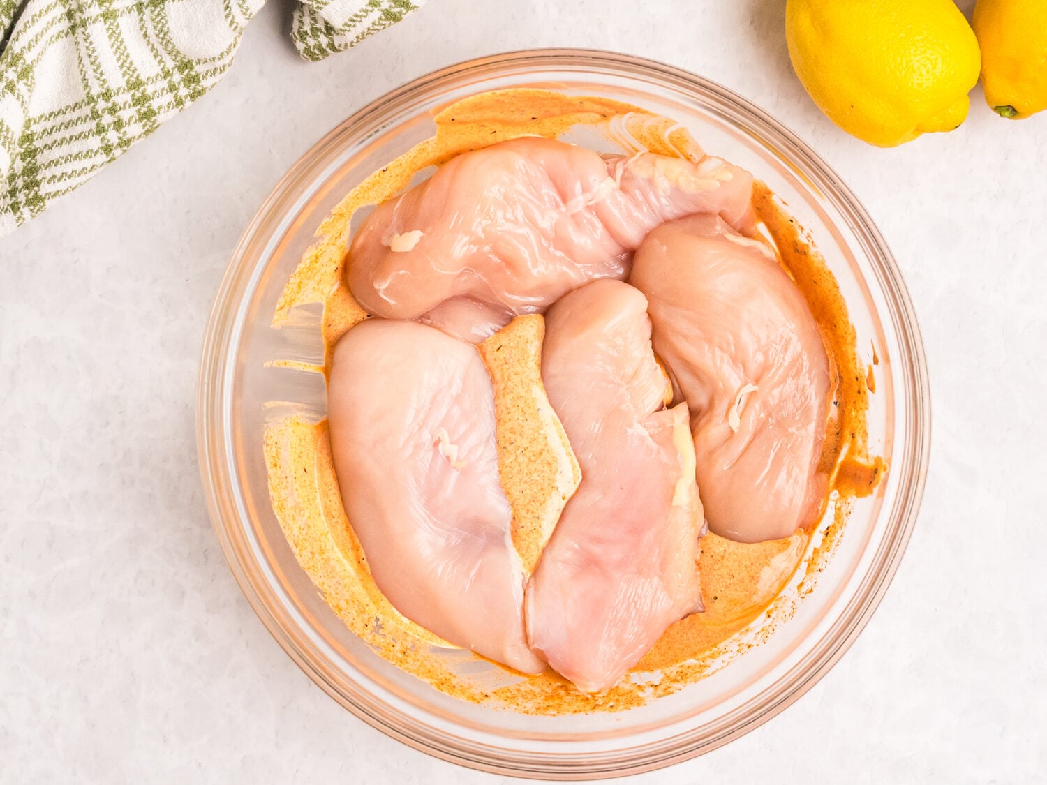 Raw chicken breasts resting in a creamy Mediterranean-style marinade inside a glass bowl, with lemons and olive oil nearby on a white countertop.
