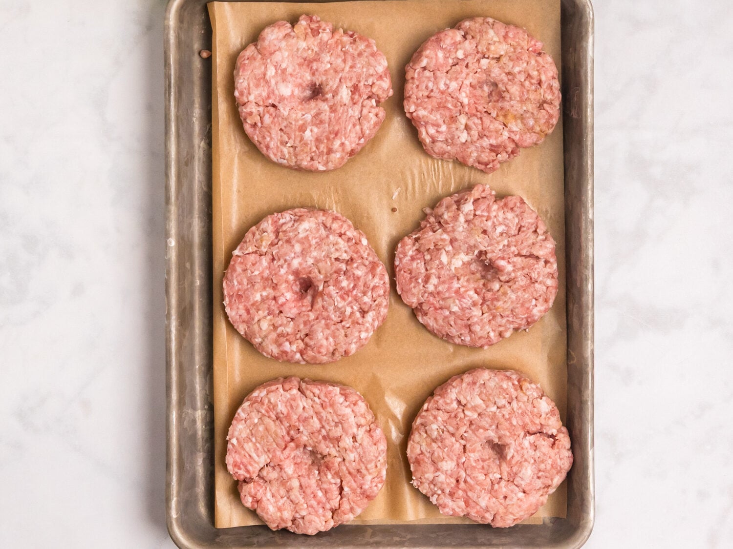 Six raw pork burger patties arranged on a parchment-lined baking sheet with lettuce, tomato slices, onion slices, and salt and pepper nearby for serving.
