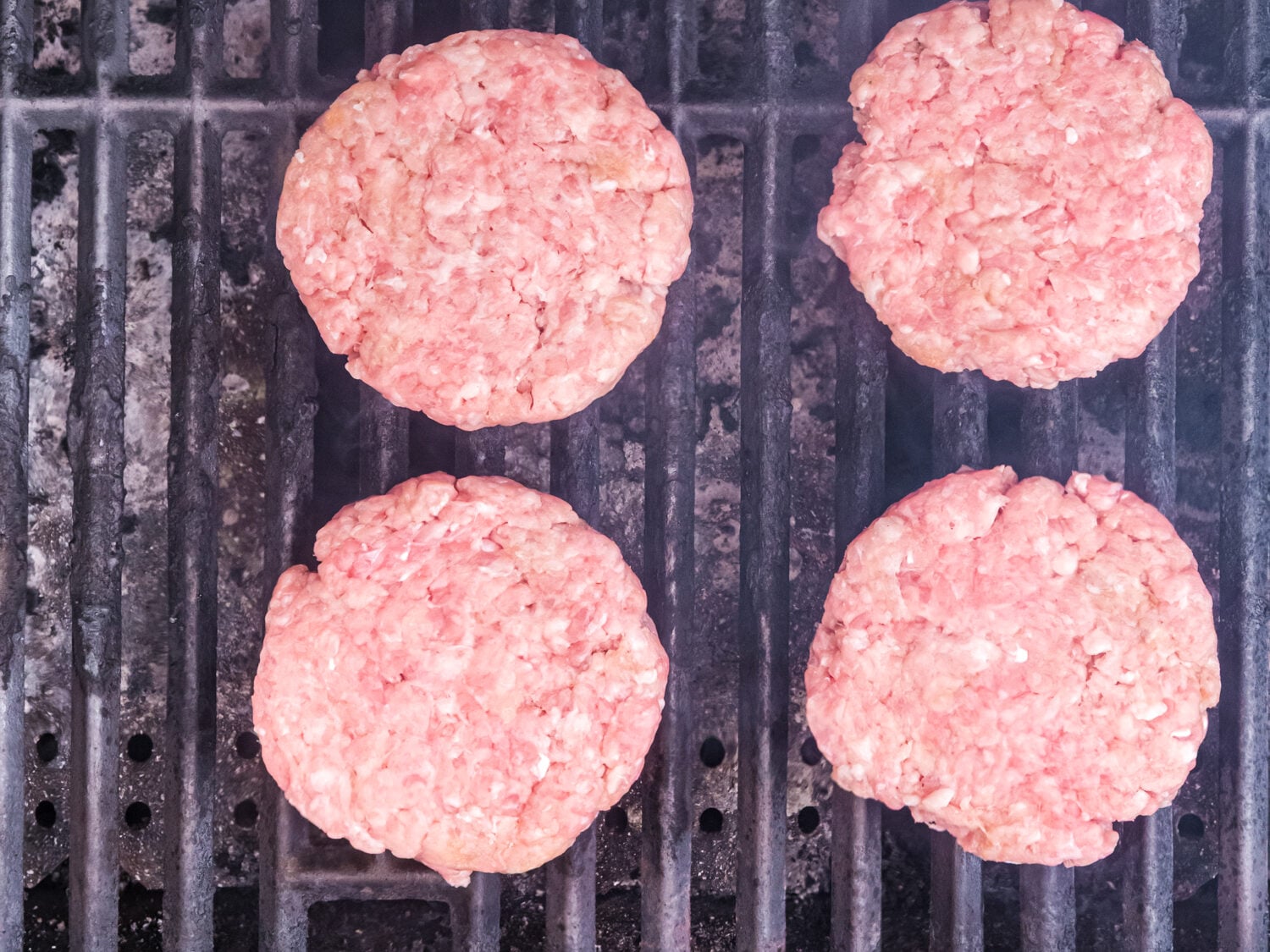 Six pork burger patties cooking on an outdoor grill, viewed from above as they begin to brown over the grates.