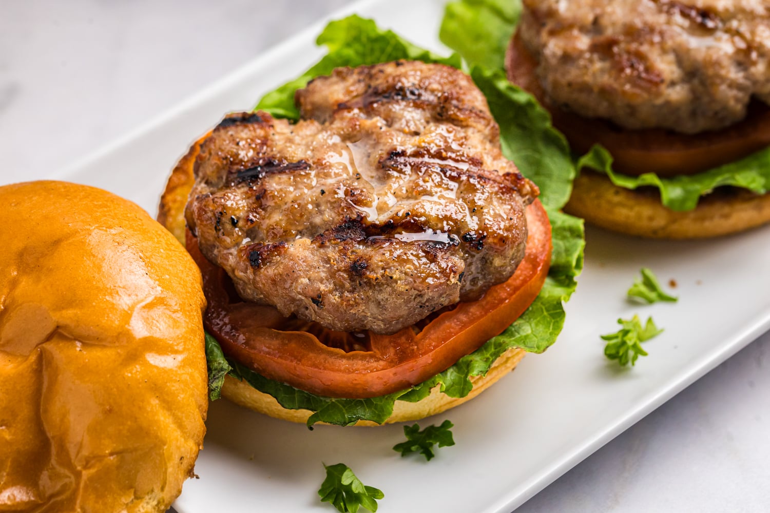 Grilled pork burgers topped with lettuce and tomato on toasted buns, served on a platter with visible grill marks and a glossy finish.