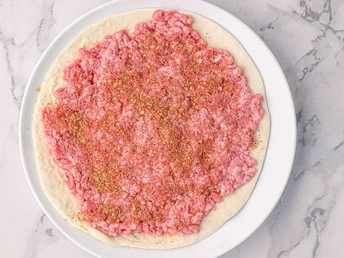 Adding beef seasoning to round beef on tortilla for Smash Burger Taco recipe