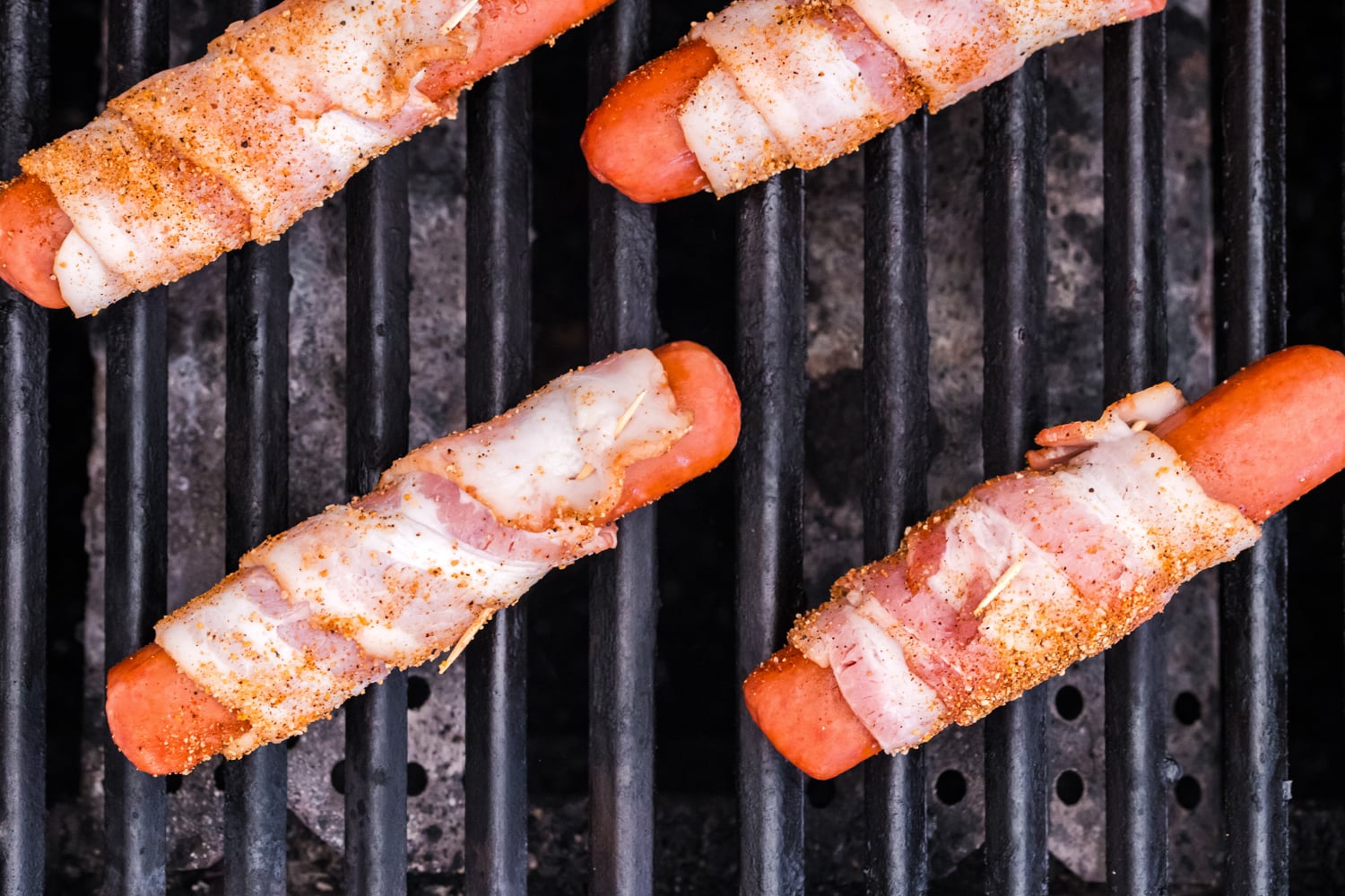 Bacon wrapped hot dogs cooking on a grill grates after being seasoned with barbecue rub.