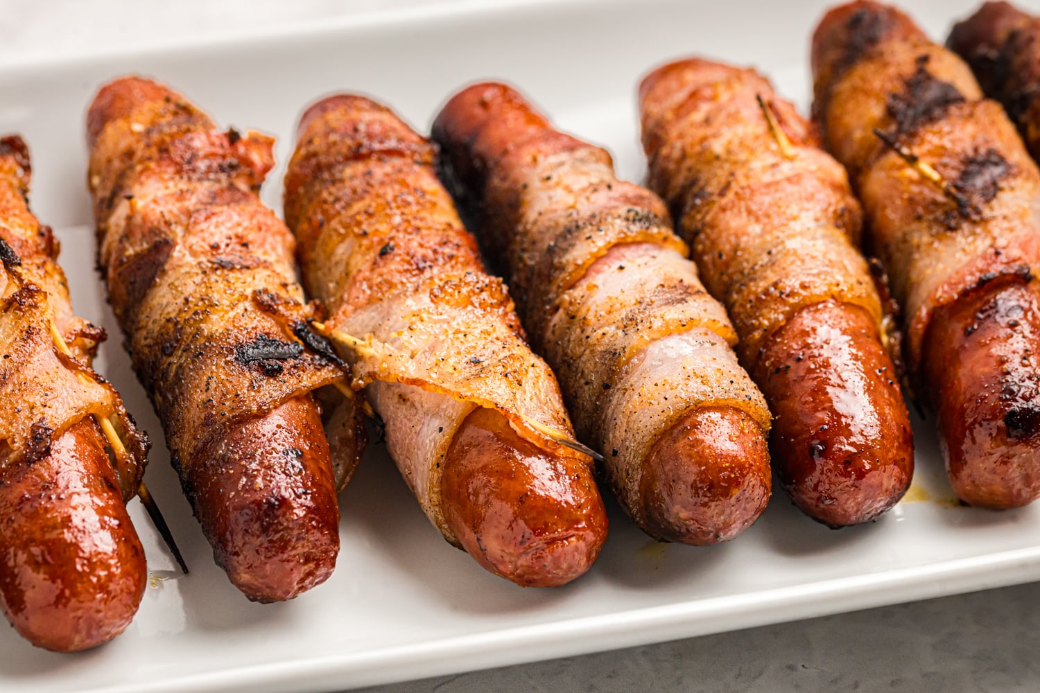 Close-up of grilled bacon wrapped hot dogs with crispy bacon served on a white platter.
