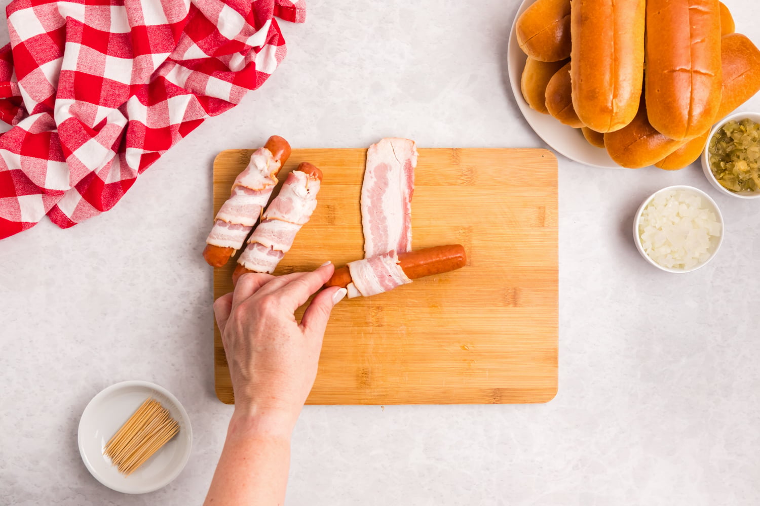 Hand wrapping hot dogs with bacon on a cutting board before securing them with toothpicks for grilling.