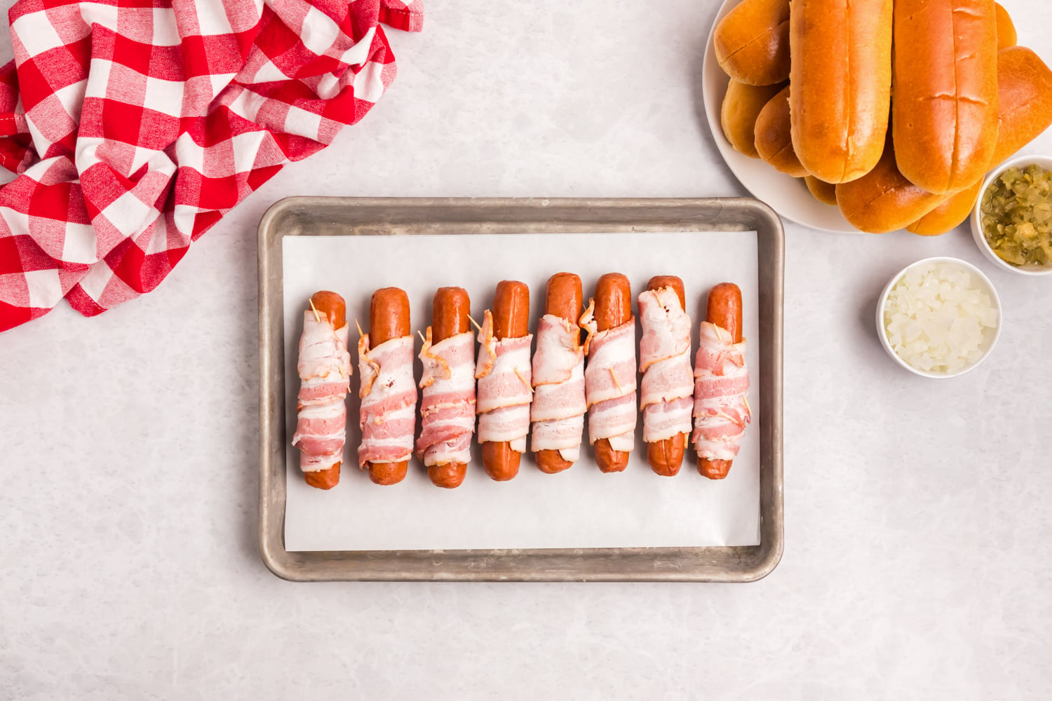 Tray of uncooked bacon wrapped hot dogs lined up on a parchment-lined baking sheet before grilling.
