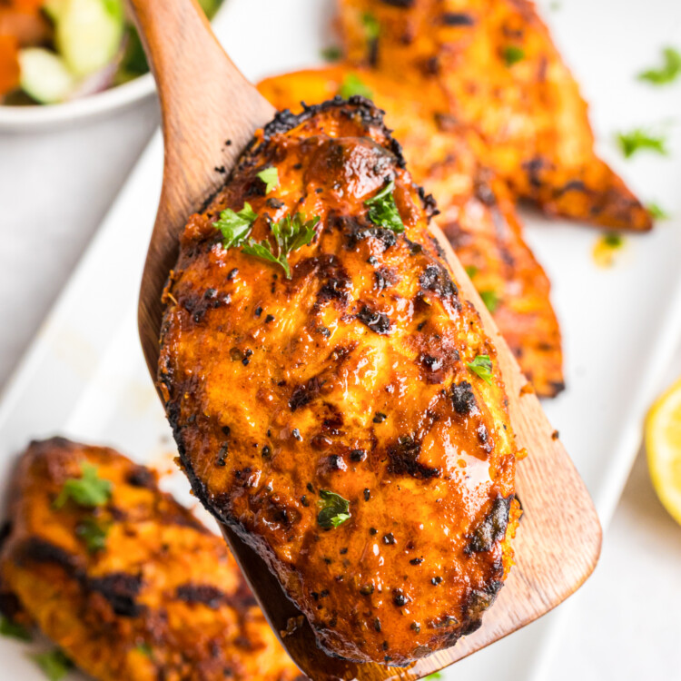 Grilled Mediterranean chicken breasts with charred edges served on a platter alongside cucumber tomato salad, fresh lemon halves, and olive oil.