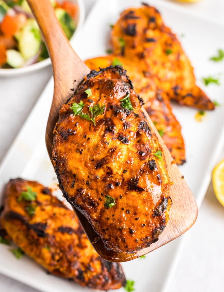 Grilled Mediterranean chicken breasts with charred edges served on a platter alongside cucumber tomato salad, fresh lemon halves, and olive oil.