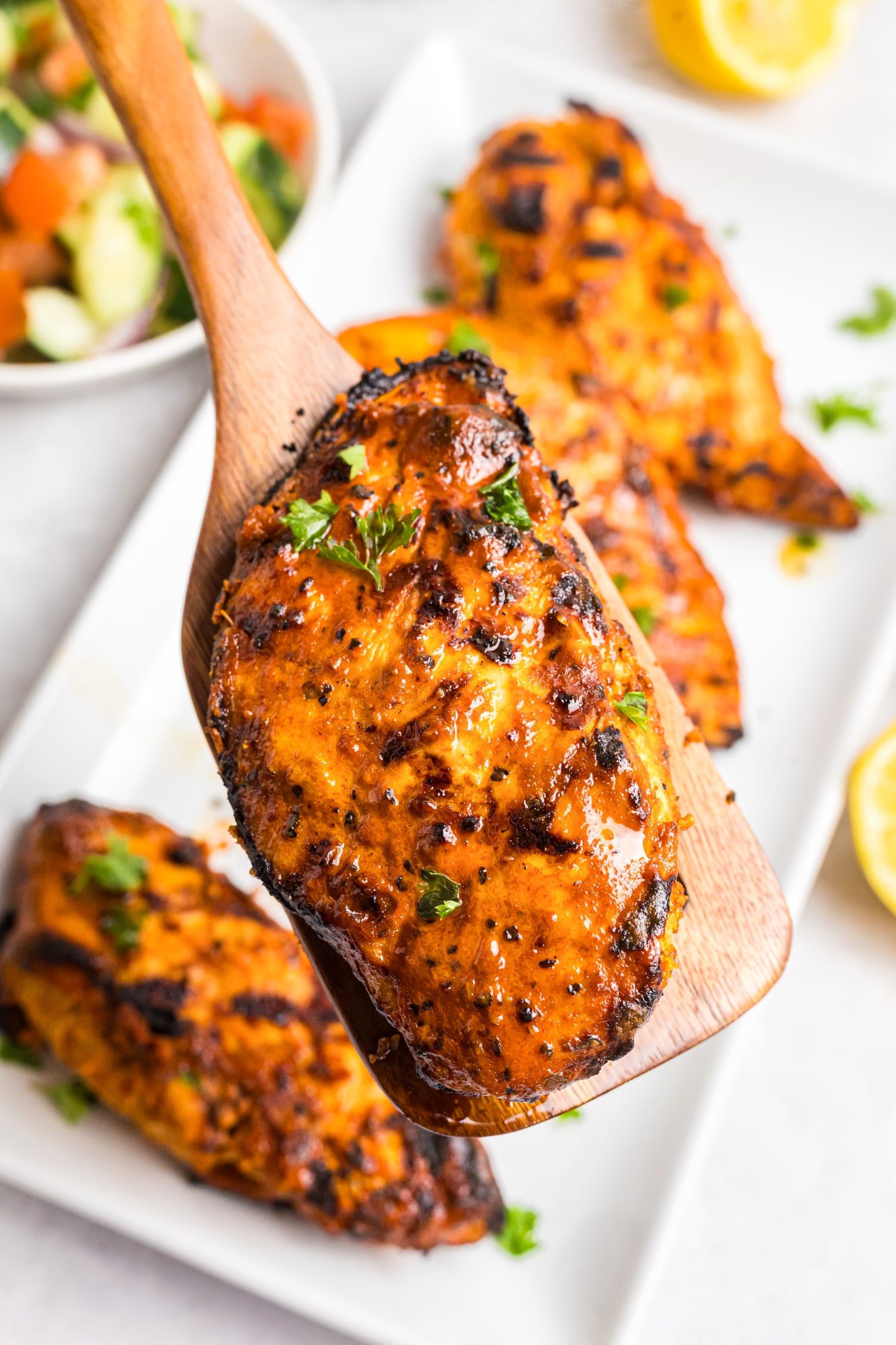 Grilled Mediterranean chicken breasts with charred edges served on a platter alongside cucumber tomato salad, fresh lemon halves, and olive oil.