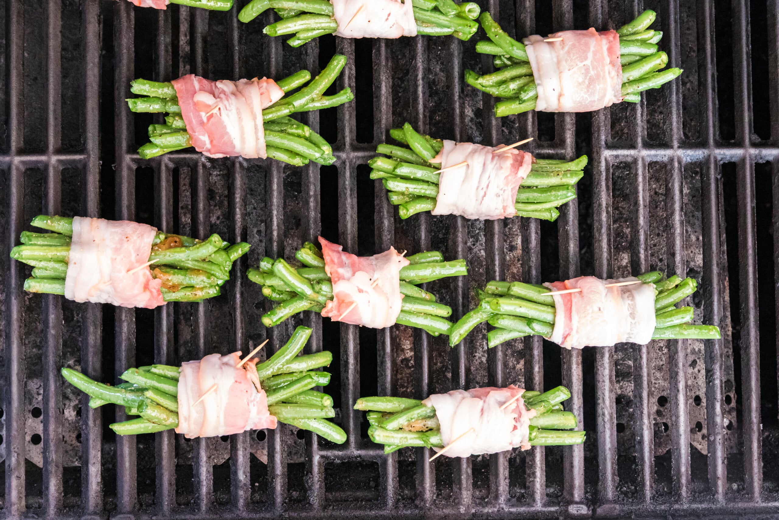 Bacon-wrapped green bean bundles secured with toothpicks and arranged on a hot outdoor grill.