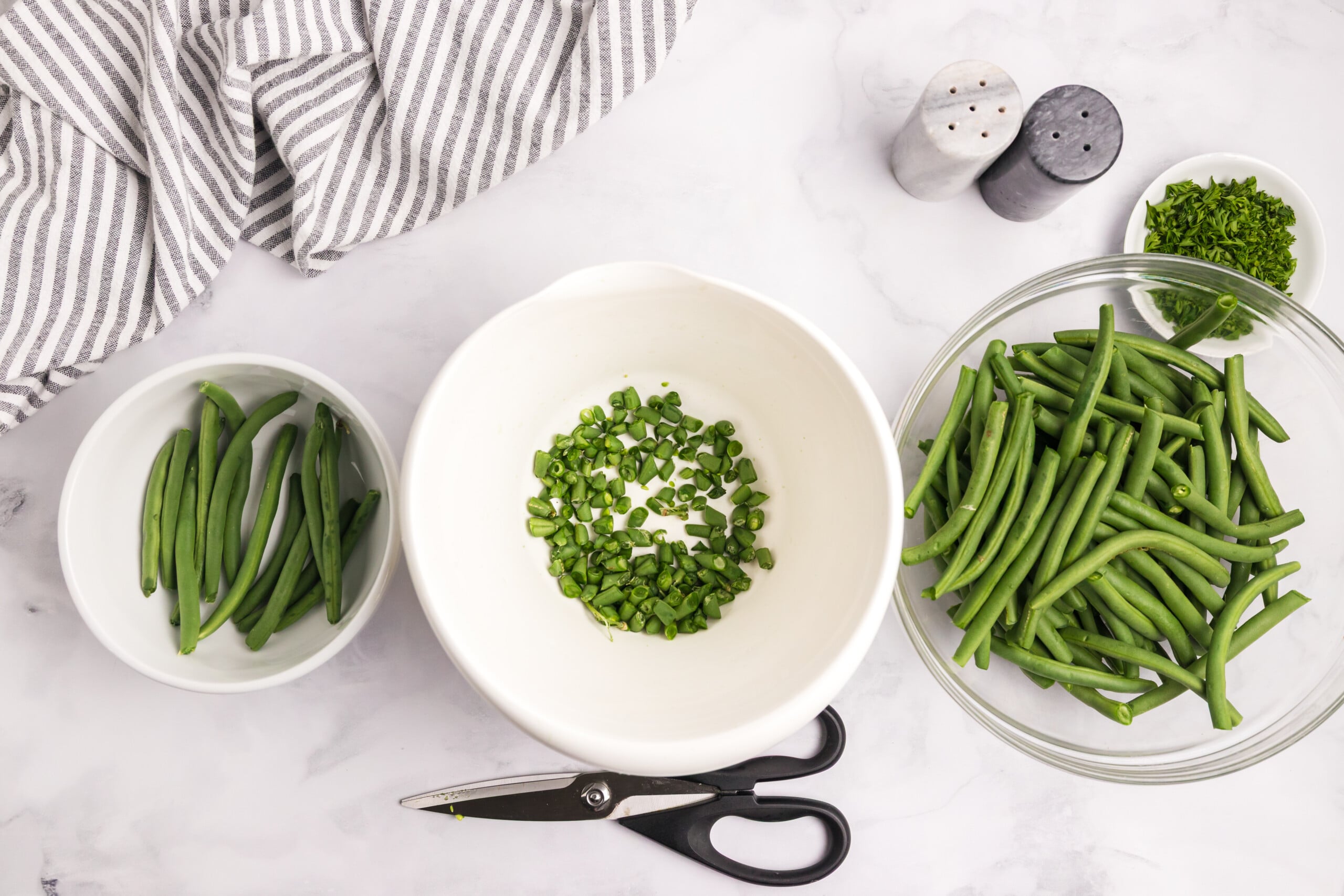 Fresh green beans being trimmed with kitchen shears, with bowls of whole beans, parsley, salt, and pepper nearby.