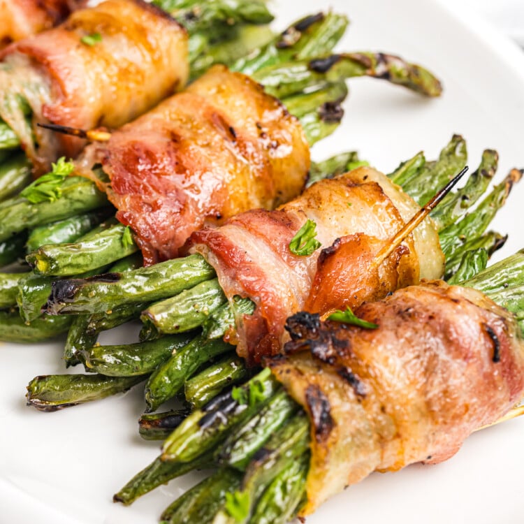 Grilled bacon-wrapped green bean bundles with crisp, browned bacon and lightly charred green beans served on a white platter.