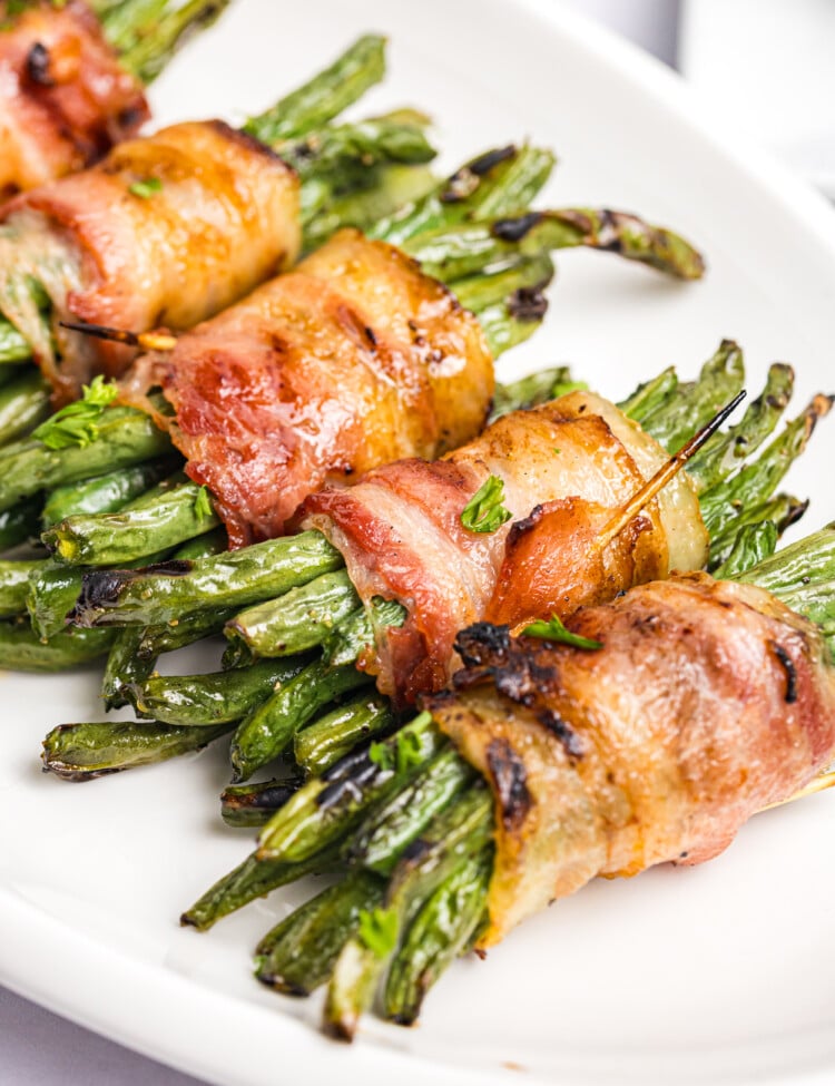 Grilled bacon-wrapped green bean bundles with crisp, browned bacon and lightly charred green beans served on a white platter.