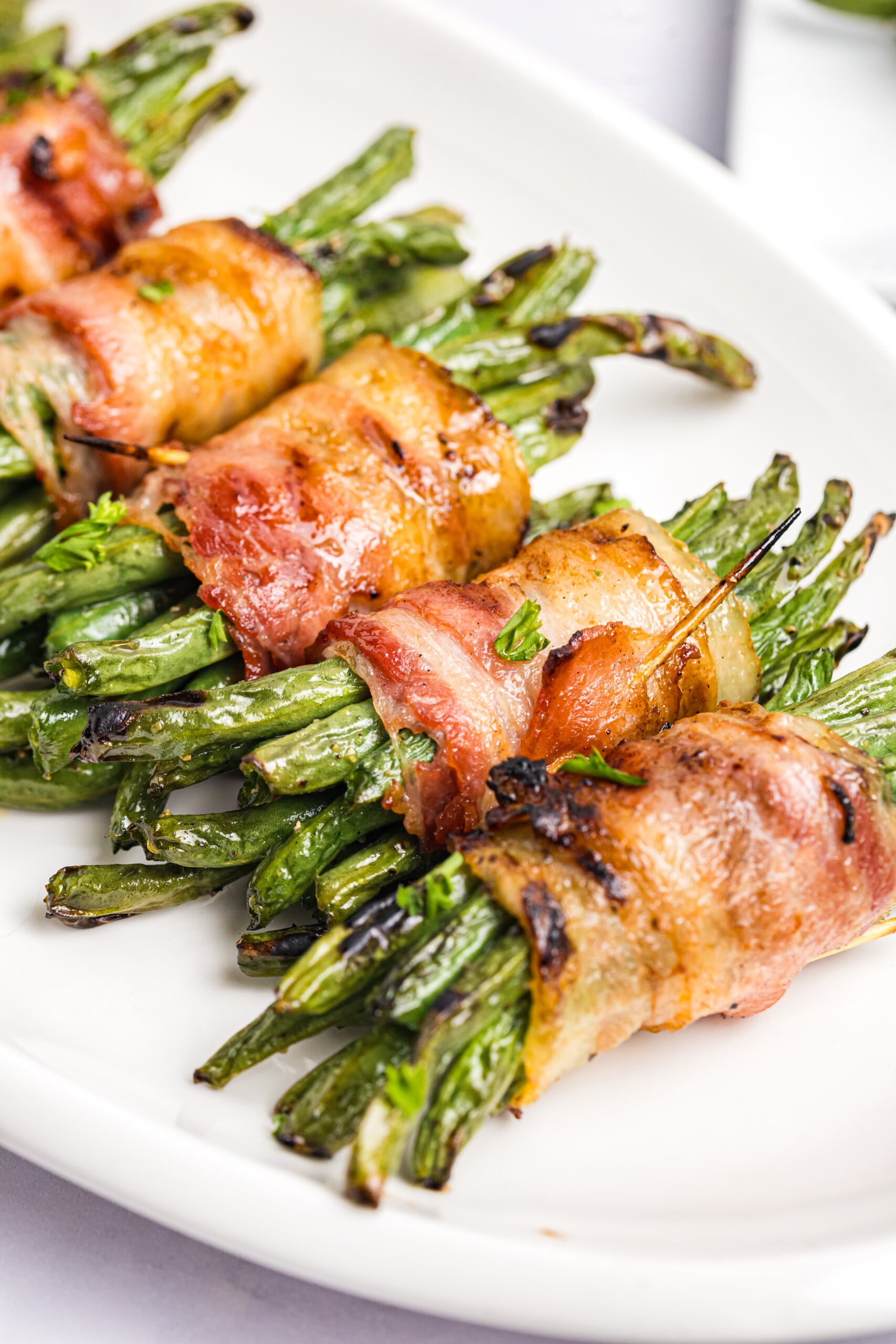 Grilled bacon-wrapped green bean bundles with crisp, browned bacon and lightly charred green beans served on a white platter.