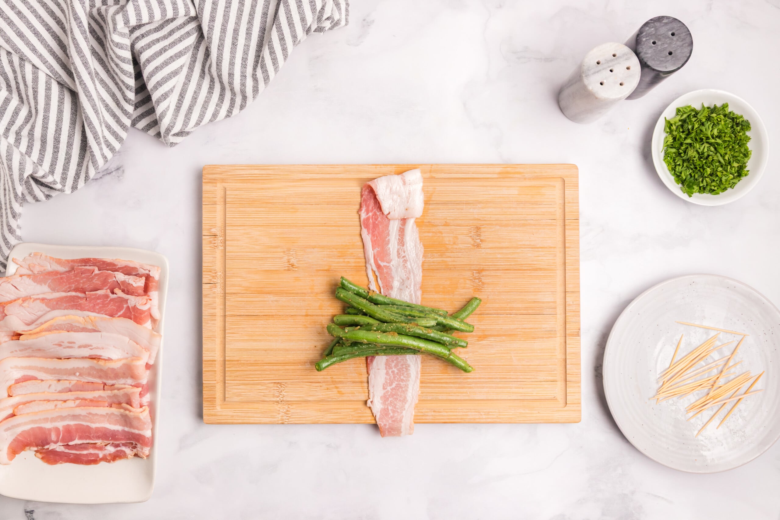 Seasoned green beans arranged on a strip of raw bacon on a cutting board, ready to be wrapped into a bundle.