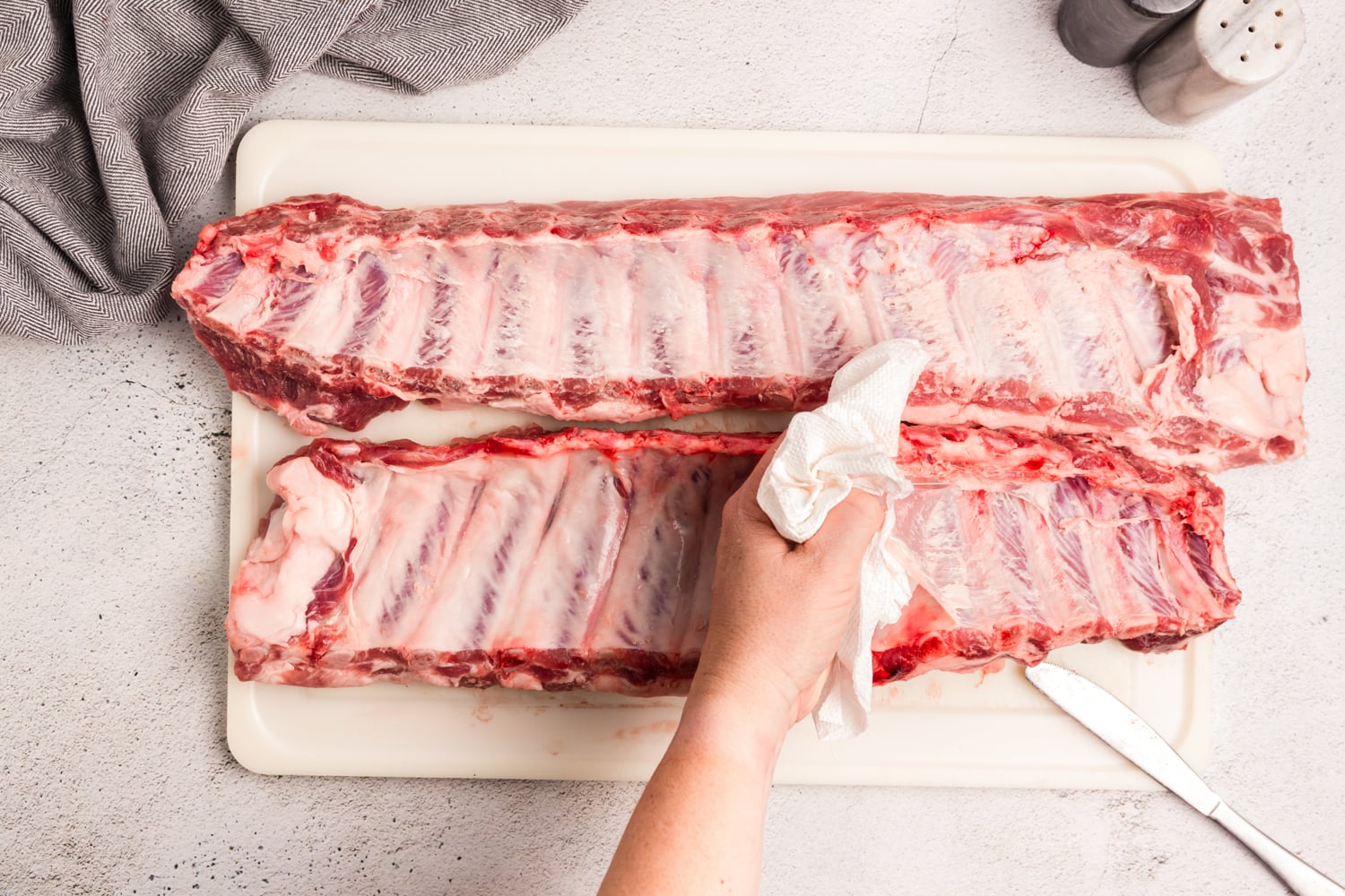 Using a paper towel to grip and remove the membrane from the back of a rack of pork ribs.