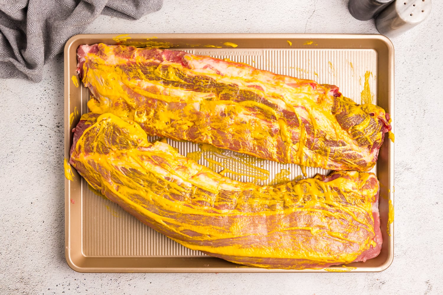 Pork ribs coated with yellow mustard on a baking sheet to help the dry rub stick before grilling.