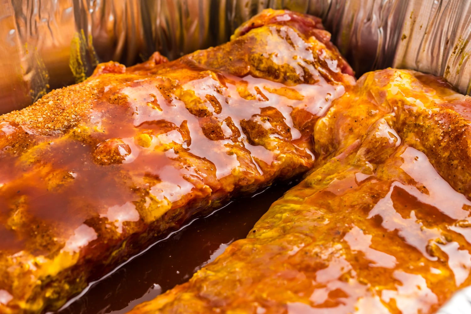 Seasoned pork ribs resting in a foil pan with barbecue sauce before cooking on the grill.