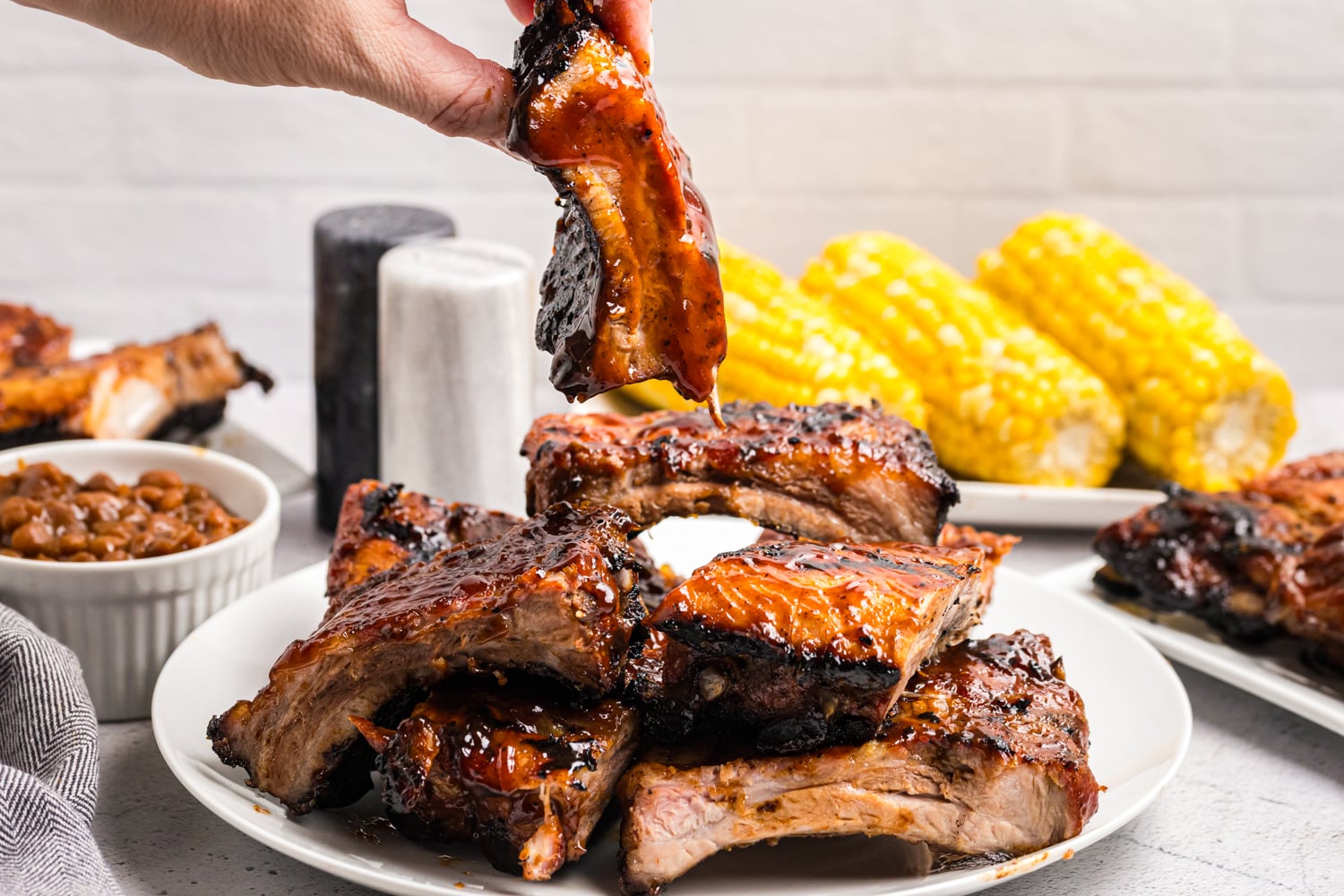 Hand lifting a sauced grilled pork rib from a platter with corn on the cob and baked beans in the background.