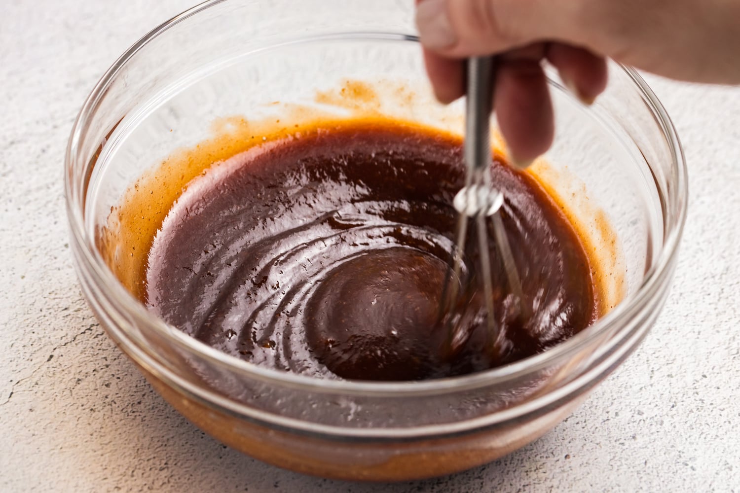 Whisking homemade barbecue sauce in a glass mixing bowl until smooth and glossy.