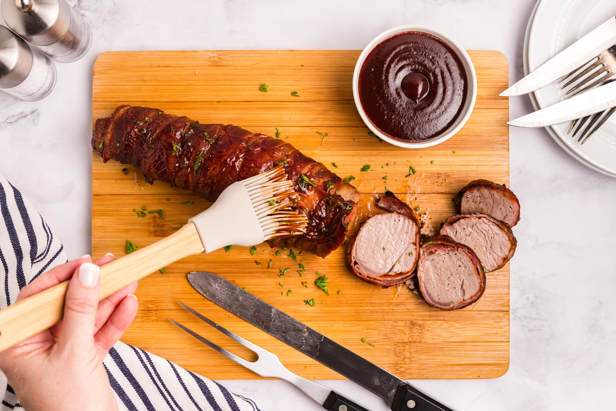 Smoked bacon-wrapped pork tenderloin being brushed with barbecue sauce, with several slices cut and ready to serve.