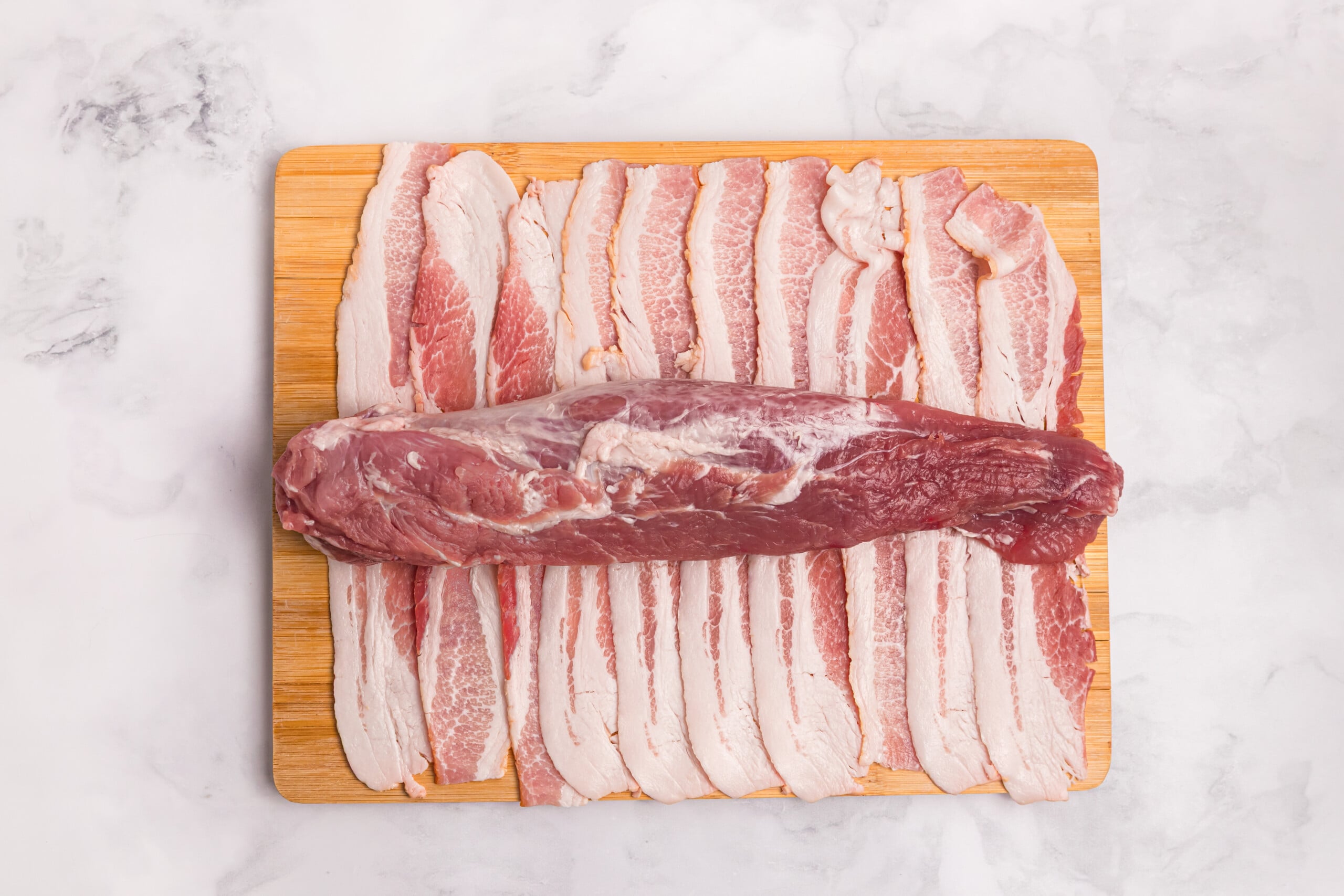 Raw pork tenderloin placed across overlapping strips of bacon on a wooden cutting board.
