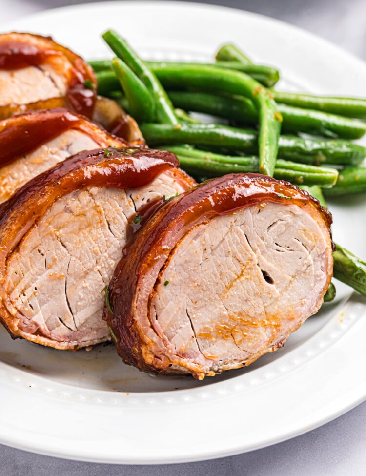 Close-up of sliced smoked bacon-wrapped pork tenderloin glazed with barbecue sauce and served with green beans.