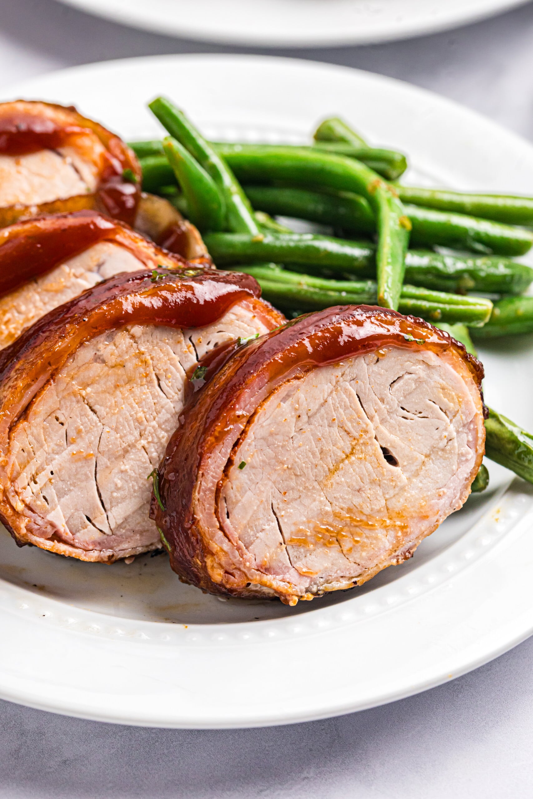 Close-up of sliced smoked bacon-wrapped pork tenderloin glazed with barbecue sauce and served with green beans.
