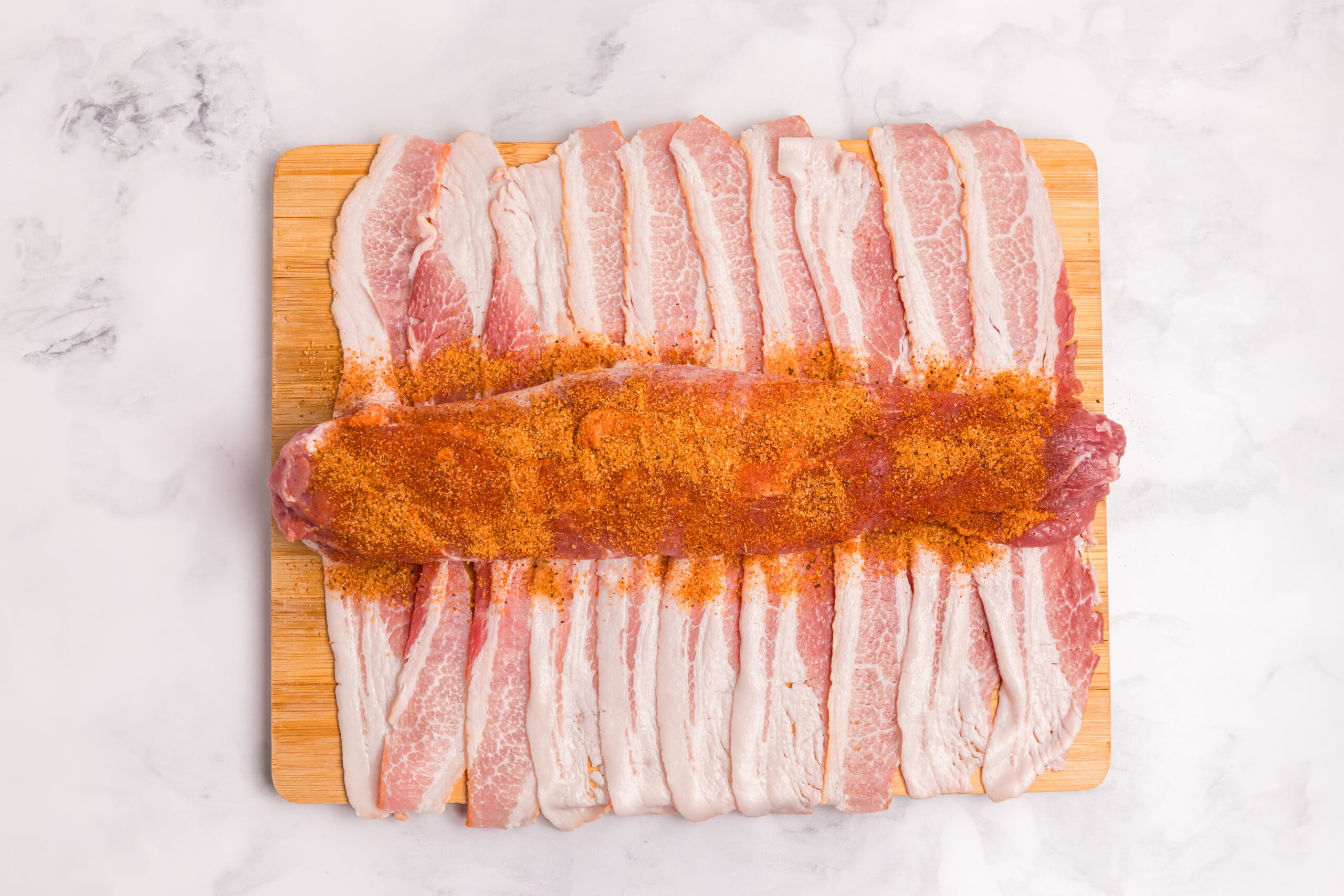 Pork tenderloin coated with a reddish-brown dry rub and positioned over strips of raw bacon.