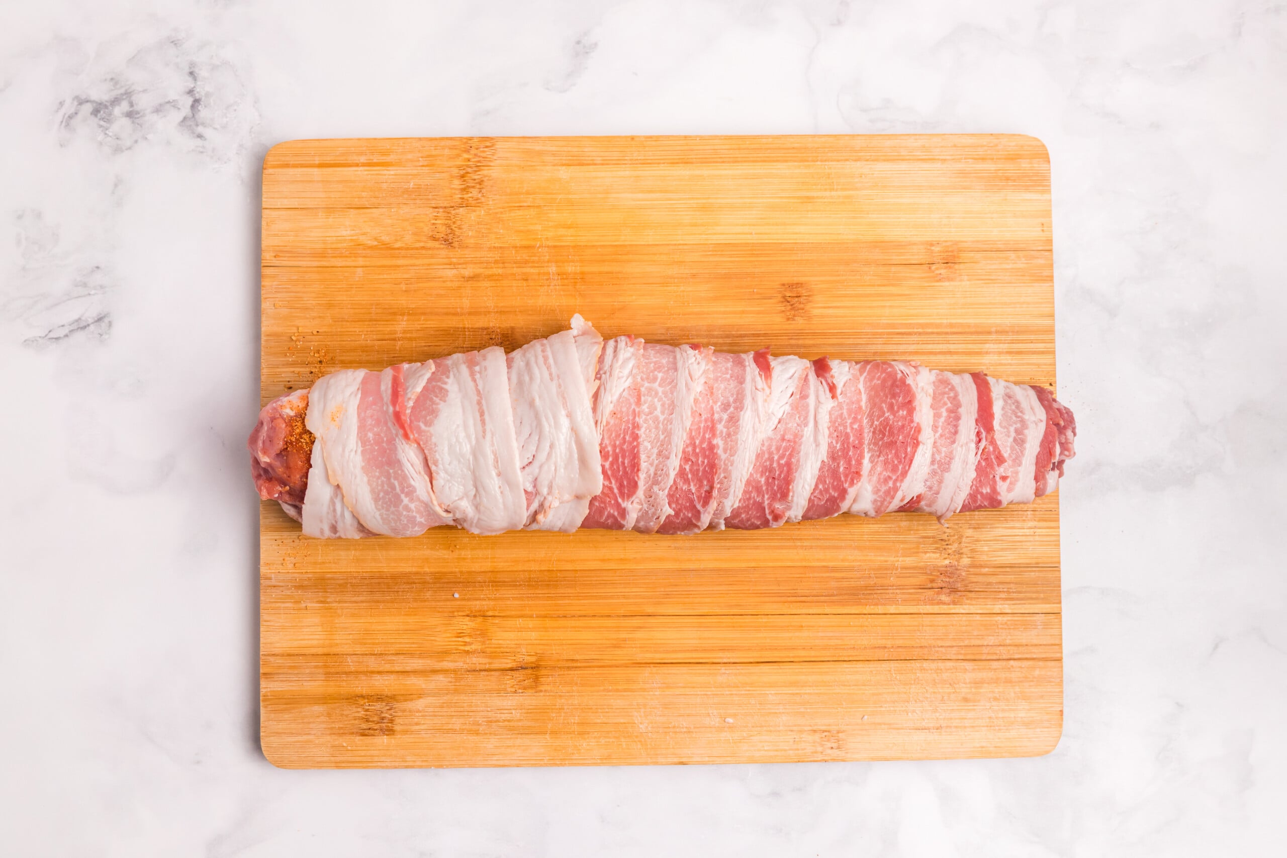Seasoned pork tenderloin completely wrapped in overlapping bacon strips on a wooden cutting board.