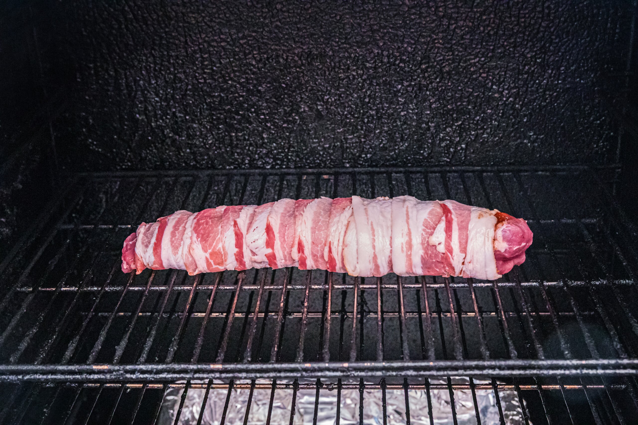 Bacon-wrapped pork tenderloin placed on the rack of a smoker, ready to cook.